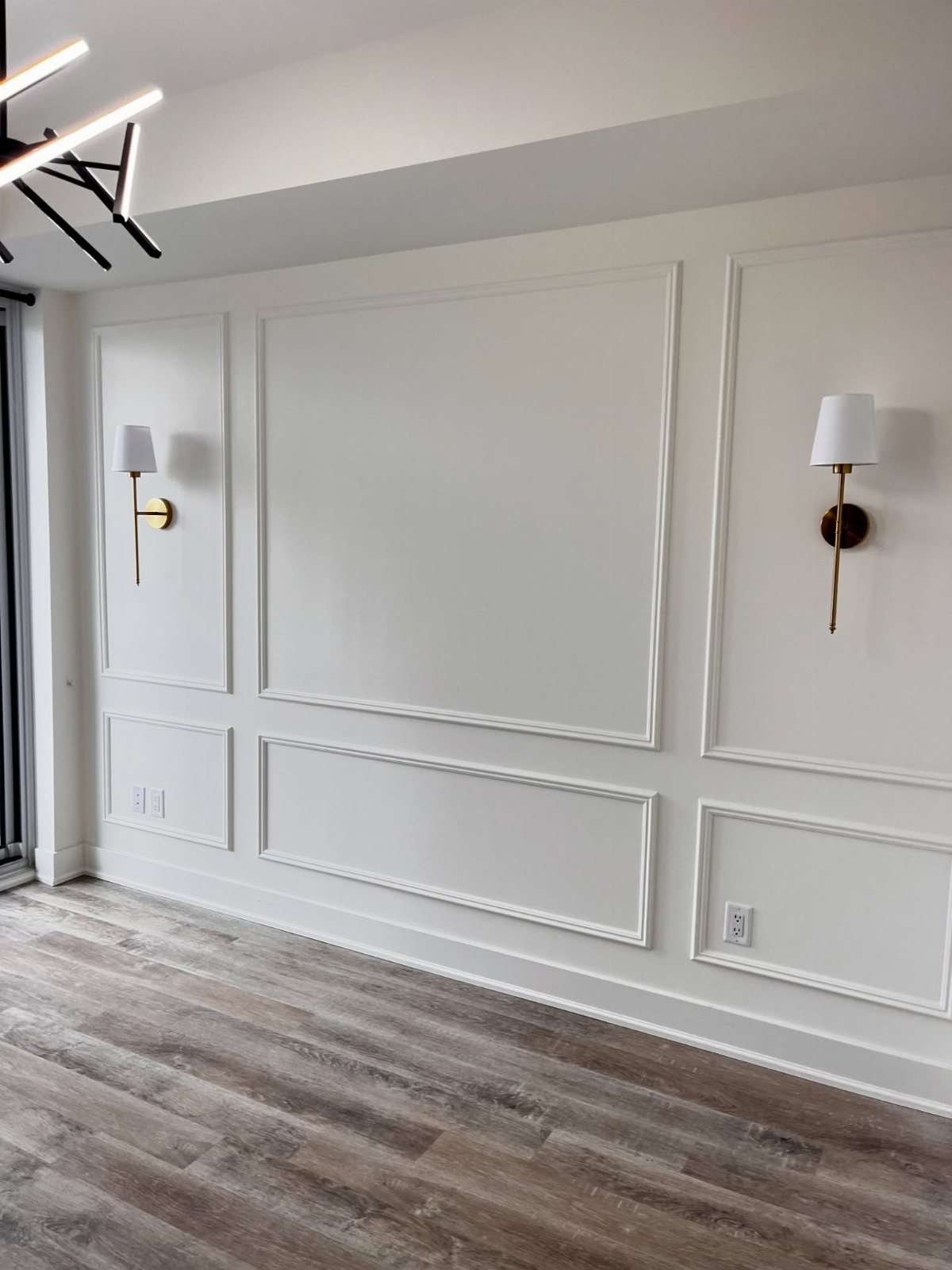 White wall with modern rectangular picture frame wainscoting and gold wall sconces above grey flooring.