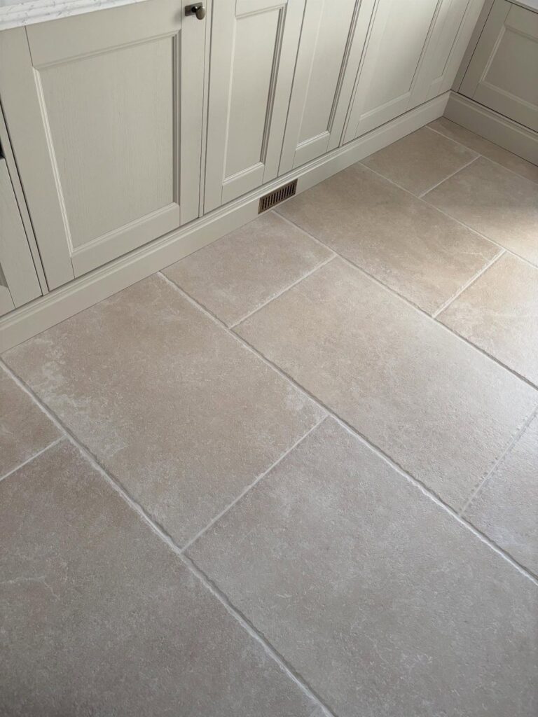 Large beige stone-effect floor tiles laid in a staggered pattern next to grey kitchen cabinets.