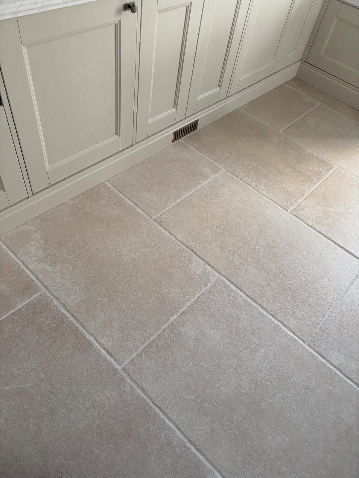 Large beige stone-effect floor tiles laid in a staggered pattern next to grey kitchen cabinets.